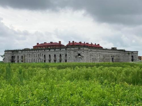Historic stone building with a red-roofed structure, surrounded by tall green grass under a cloudy sky.