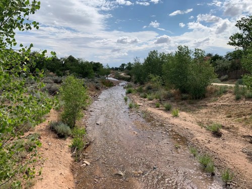 A winding creek flows through a sandy landscape, surrounded by green trees and shrubs under a partly cloudy sky.