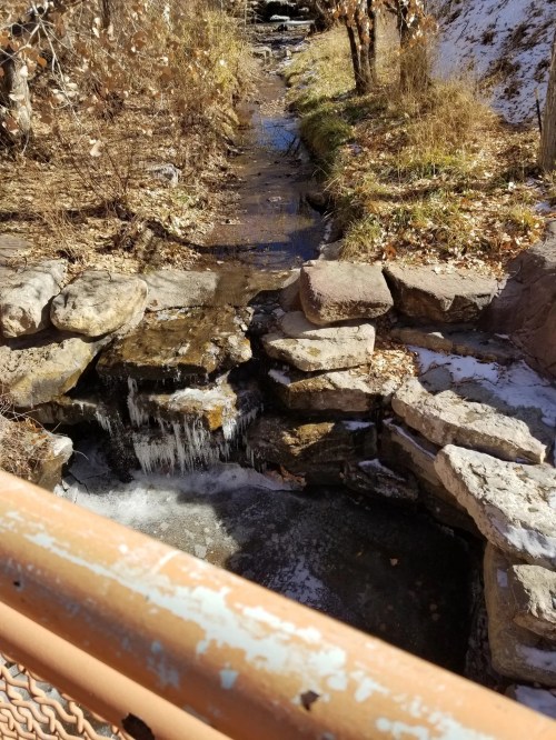 A small stream flows over rocks, surrounded by grass and trees, with a hint of snow on the ground.