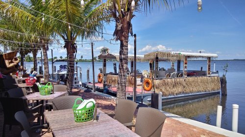 A waterfront dining area with palm trees, tables, and a thatched-roof boat docked nearby under a clear blue sky.