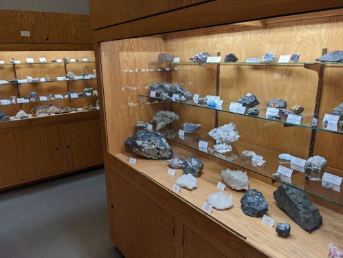 Display of various minerals and rocks in glass cases, with labels, in a well-lit museum setting.