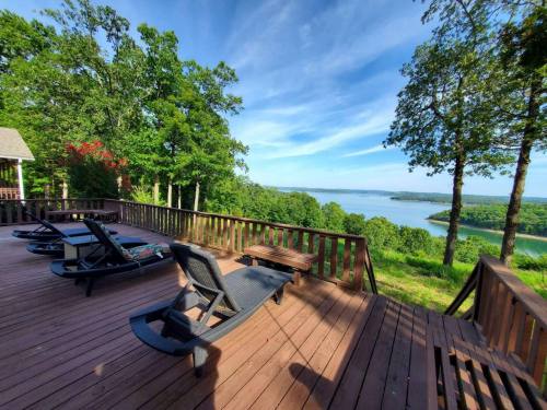 A scenic view from a wooden deck overlooking a lake, surrounded by trees and blue skies. Lounge chairs are arranged on the deck.