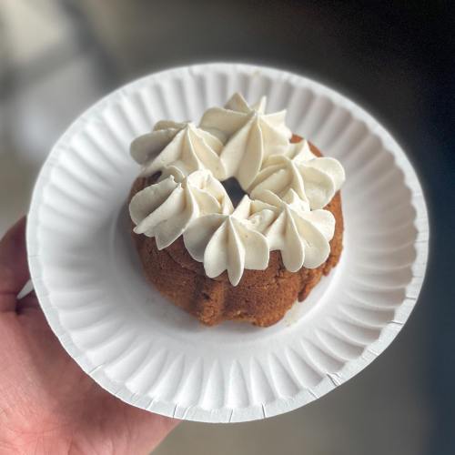 A hand holding a small bundt cake topped with swirls of white frosting on a paper plate.