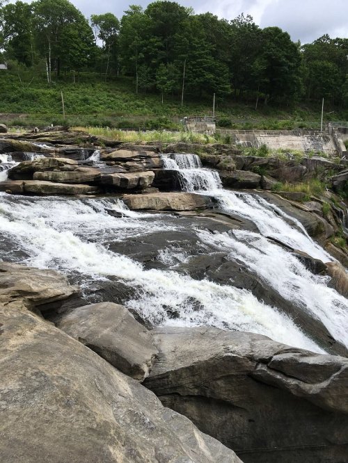 A cascading waterfall flows over rocky terrain, surrounded by lush greenery and trees in the background.