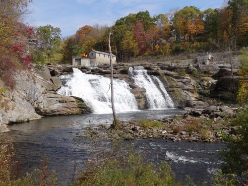 A scenic view of a waterfall cascading over rocks, surrounded by autumn foliage and a calm river.