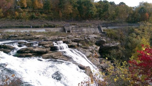 A scenic view of a waterfall cascading over rocks, surrounded by autumn foliage and a dam in the background.