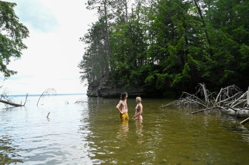 Two people wade in shallow water surrounded by trees and rocky cliffs on a calm day.