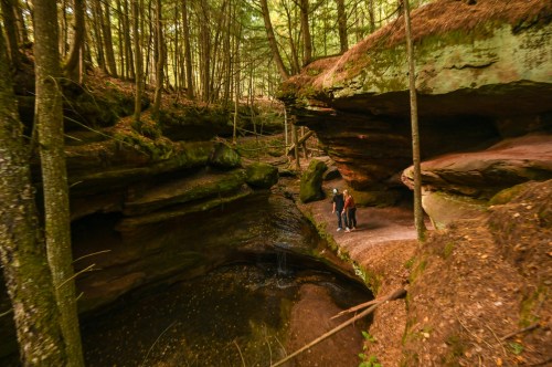 Two people stand by a serene, wooded area with rock formations and a small pond surrounded by trees.