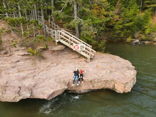 Two people sit on a rocky outcrop by a river, with a wooden staircase and trees in the background.