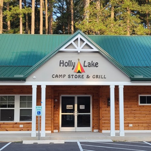 Exterior of Holly Lake Camp Store & Grill, featuring a wooden facade, green roof, and surrounded by trees.