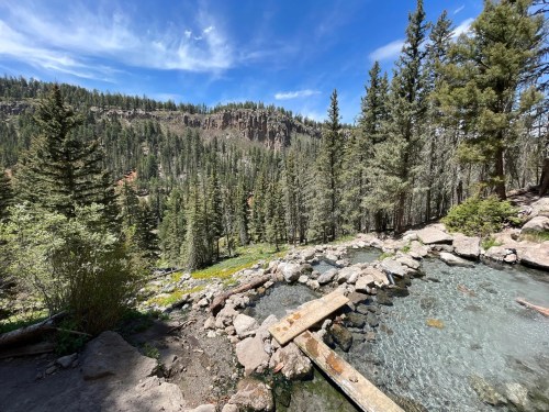 A serene natural hot spring surrounded by lush trees and rocky hills under a clear blue sky.