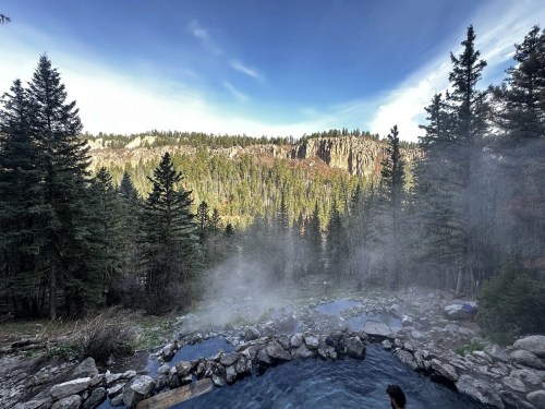 A serene hot spring surrounded by lush trees and a misty landscape, with mountains in the background under a clear sky.