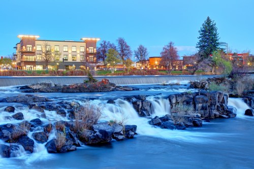 A serene river flows over rocky terrain, with a charming building and trees in the background under a twilight sky.
