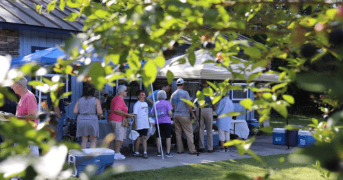 A group of people gather under tents outside, enjoying a sunny day at a community event. Green foliage frames the scene.