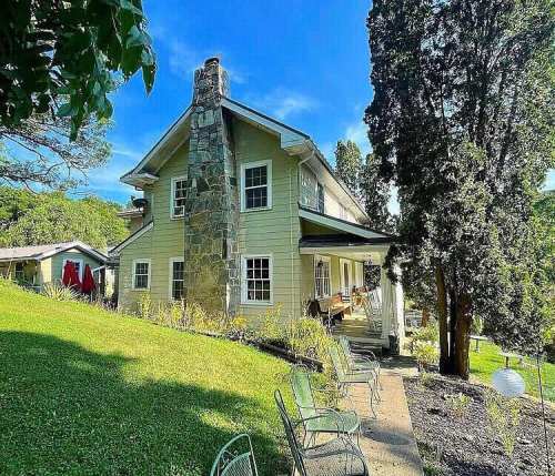 A charming two-story house with a stone chimney, surrounded by greenery and outdoor seating on a sunny day.