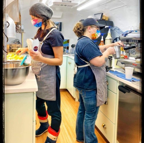 Two women in striped aprons and colorful masks prepare food and drinks in a bright kitchen.