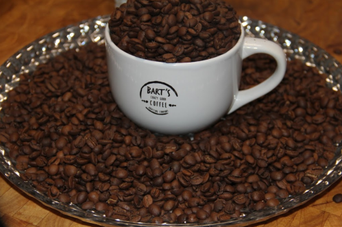 A white coffee cup filled with coffee beans, surrounded by a pile of beans on a silver tray.