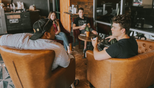 Two young men sit in armchairs, engaged in conversation, while two others relax in the background at a cozy café.