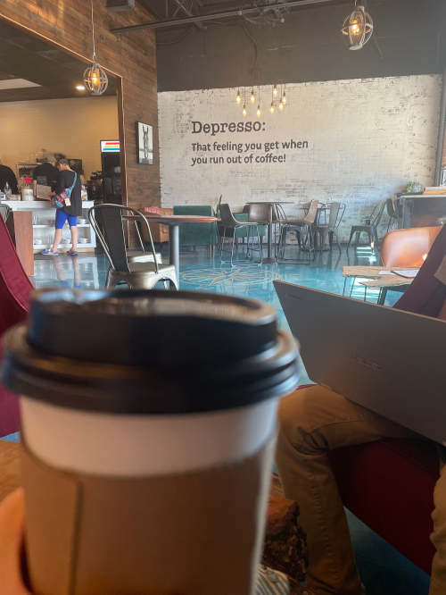 A coffee cup in the foreground with a cozy café setting and a humorous wall quote about coffee in the background.