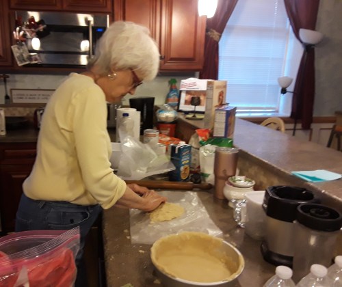 An older woman in a yellow sweater prepares dough on a kitchen counter filled with various cooking ingredients.