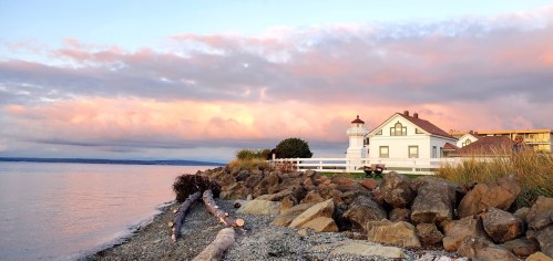 A scenic view of a lighthouse by the water, with a house and colorful clouds at sunset.