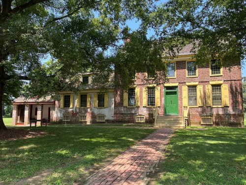 Historic brick building with yellow shutters and a green door, surrounded by trees and a grassy area.