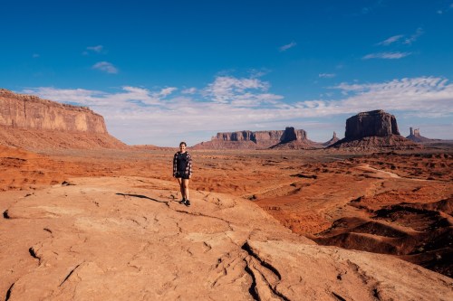 A person stands on rocky terrain in a vast desert landscape with red cliffs and blue skies in the background.