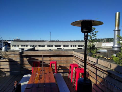 Rooftop view with wooden seating, red stools, and a heater, overlooking a building and clear blue sky.