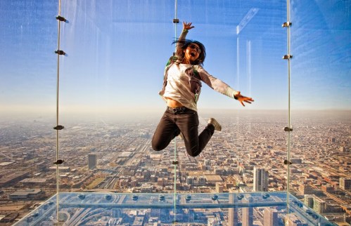 A person joyfully jumping inside a glass observation box high above a city skyline.