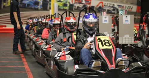 A line of go-karts with drivers wearing helmets, ready to race on an indoor track.