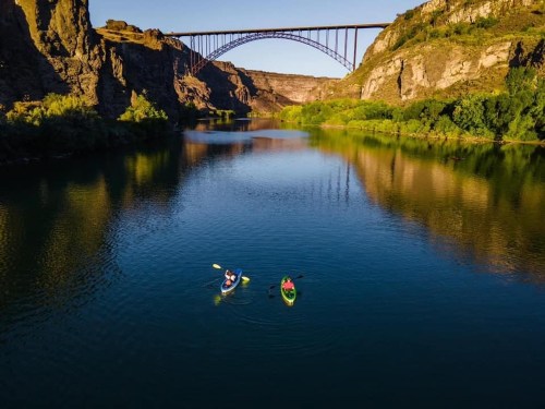 Two kayakers paddle on a calm river beneath a large bridge, surrounded by rocky cliffs and lush greenery.