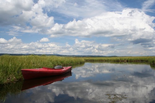 A red kayak rests on calm water, reflecting fluffy clouds and green grass under a blue sky.