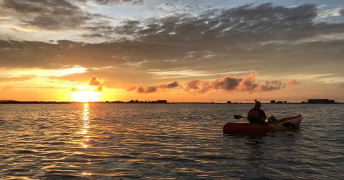 A person kayaking on calm water at sunset, with vibrant orange and pink clouds in the sky.