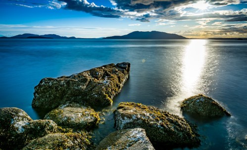 A serene seascape featuring rocky formations, calm waters, and a sunset illuminating distant mountains.