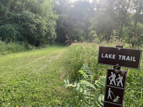 A grassy path leads into a wooded area, marked by a sign for the Lake Trail with icons for walking and pets.