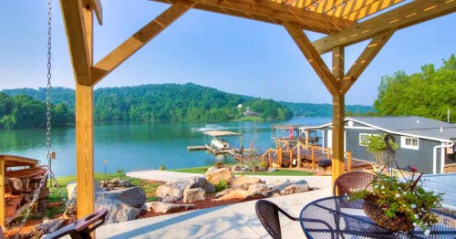 A scenic view of a lake surrounded by lush green hills, framed by a wooden furnished deck at a lakefront Airbnb in Tennessee.