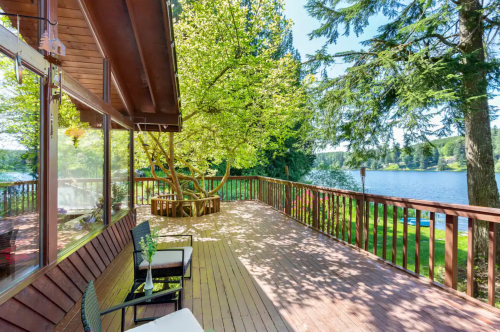 A sunny deck with chairs overlooks a serene lake surrounded by trees.