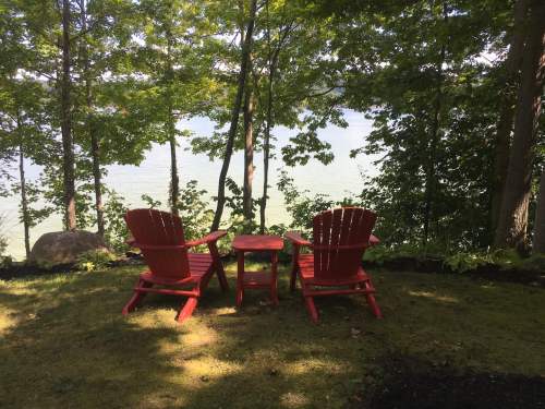 Two red Adirondack chairs and a small table facing a serene lake, surrounded by lush green trees.