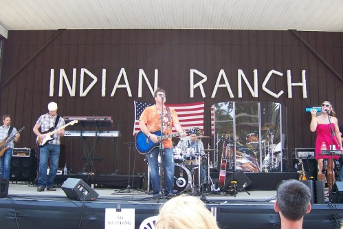 A band performs on stage at Indian Ranch, with an American flag in the background and an audience in front.
