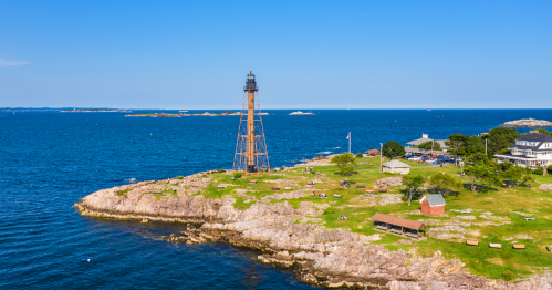 A scenic view of a lighthouse on rocky land by the ocean, with clear blue skies and distant islands.