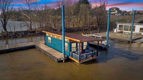 A colorful houseboat on a dock, surrounded by water and trees, with a sunset sky in the background.