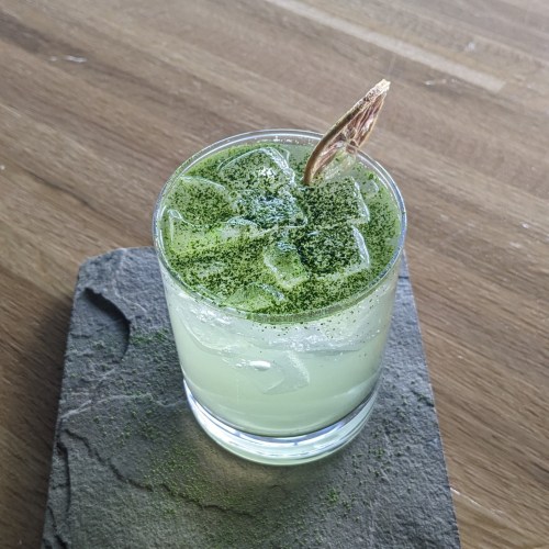 A refreshing green cocktail with ice, garnished with a slice of lemon grass, served on a slate coaster.