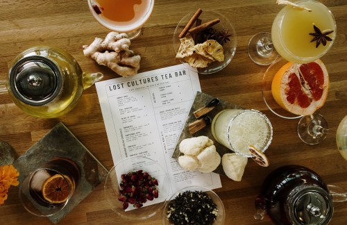 A variety of colorful drinks and tea ingredients arranged on a wooden table, with a menu for "Lost Cultures Tea Bar."
