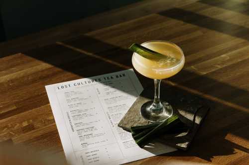 A cocktail in a coupe glass sits on a wooden table next to a menu and fresh green leaves.