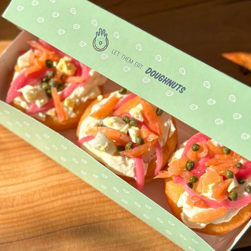 A box of three gourmet doughnuts topped with cream, pickled vegetables, and capers, set on a wooden surface.