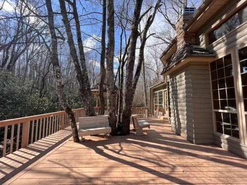 A wooden deck with two benches, surrounded by trees and a house, under a blue sky with scattered clouds.