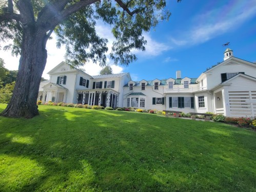 A large white house with multiple gables, surrounded by green grass and colorful flower beds under a blue sky.