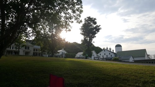 A serene rural scene with a grassy area, farm buildings, and a setting sun behind trees.