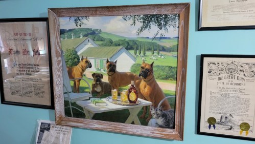 A framed painting of dogs and a cat at a table outdoors, surrounded by green fields and a farmhouse in the background.