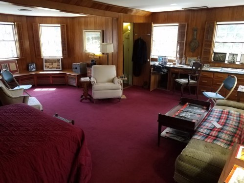 Cozy room with wooden walls, a red carpet, a bed, armchair, desk, and windows, featuring vintage decor and furniture.